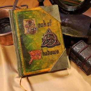 May include: A green leather-bound journal with a decorative embossed cover. The cover features the text "Book of Shadows" and several intricate designs, including a Celtic knot and a stylized floral motif.