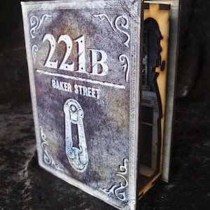 Pode incluir: Uma caixa de madeira em forma de livro com um compartimento oculto. A frente da caixa &eacute; decorada com um visual desgastado e envelhecido e apresenta o texto "221B Baker Street".