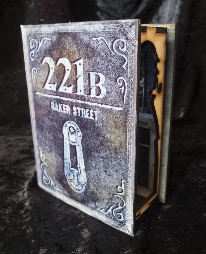 Pode incluir: Uma caixa de madeira em forma de livro com um compartimento oculto. A frente da caixa &eacute; decorada com um visual desgastado e envelhecido e apresenta o texto "221B Baker Street".
