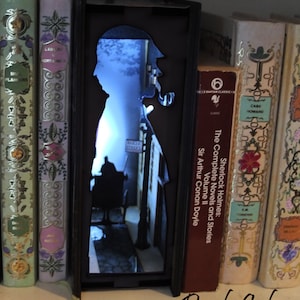 Pode incluir: Um separador de livros preto com uma silhueta de Sherlock Holmes e uma cena de rua. O separador de livros &eacute; iluminado por tr&aacute;s, criando um efeito brilhante. O separador de livros est&aacute; em uma estante com outros livros.