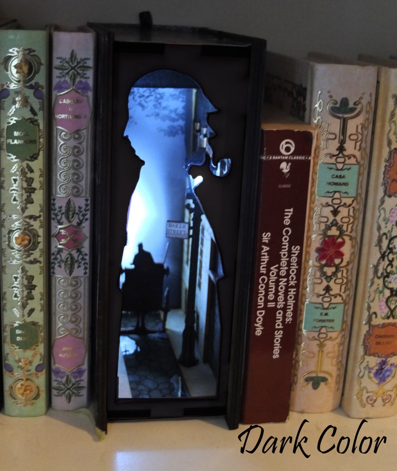 Pode incluir: Um separador de livros preto com uma silhueta de Sherlock Holmes e uma cena de rua. O separador de livros &eacute; iluminado por tr&aacute;s, criando um efeito brilhante. O separador de livros est&aacute; em uma estante com outros livros.