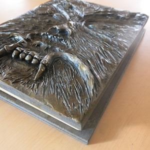 Puede incluir: Una caja rectangular de color marr&oacute;n oscuro, texturizada, con una imagen esculpida en relieve de una criatura que gru&ntilde;e con dientes afilados. La cara de la criatura est&aacute; detallada con dientes afilados y una frente arrugada.