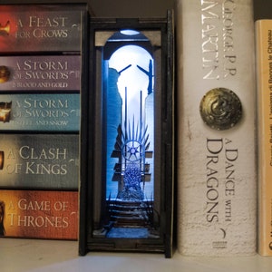 Könnte beinhalten: Ein Buchstütze mit einer Miniatur-Nachbildung des Eisernen Throns aus der Fernsehserie Game of Thrones. Der Thron ist aus Metall und hat eine schwarze Oberfläche. Die Buchstütze ist von hinten beleuchtet und erzeugt einen blauen Schein. Die Buchstütze wird zwischen zwei Büchern aus der Game of Thrones-Serie, "A Dance with Dragons" und "A Game of Thrones", angezeigt.