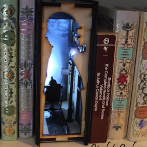 Pode incluir: Uma luz noturna de madeira em forma de livro com uma silhueta de Sherlock Holmes e uma cena de rua. A luz &eacute; iluminada com uma luz branca.