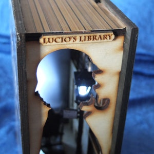 Pode incluir: Uma caixa de madeira em forma de livro com um recorte de duas silhuetas. A caixa tem uma placa que diz "Lucio's Library".