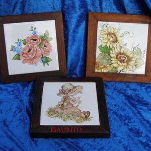 May include: Three framed ceramic tiles with painted floral and character designs. The first tile features pink roses and blue forget-me-nots. The second tile features yellow sunflowers. The third tile features a girl with a straw hat and a brown bunny.