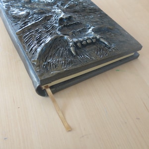 Puede incluir: Un diario de cuero gris oscuro con un dise&ntilde;o de cabeza de lobo en relieve detallado en la portada. La boca del lobo est&aacute; abierta, revelando dientes afilados. Un marcap&aacute;ginas de cinta marr&oacute;n est&aacute; unido a la encuadernaci&oacute;n.