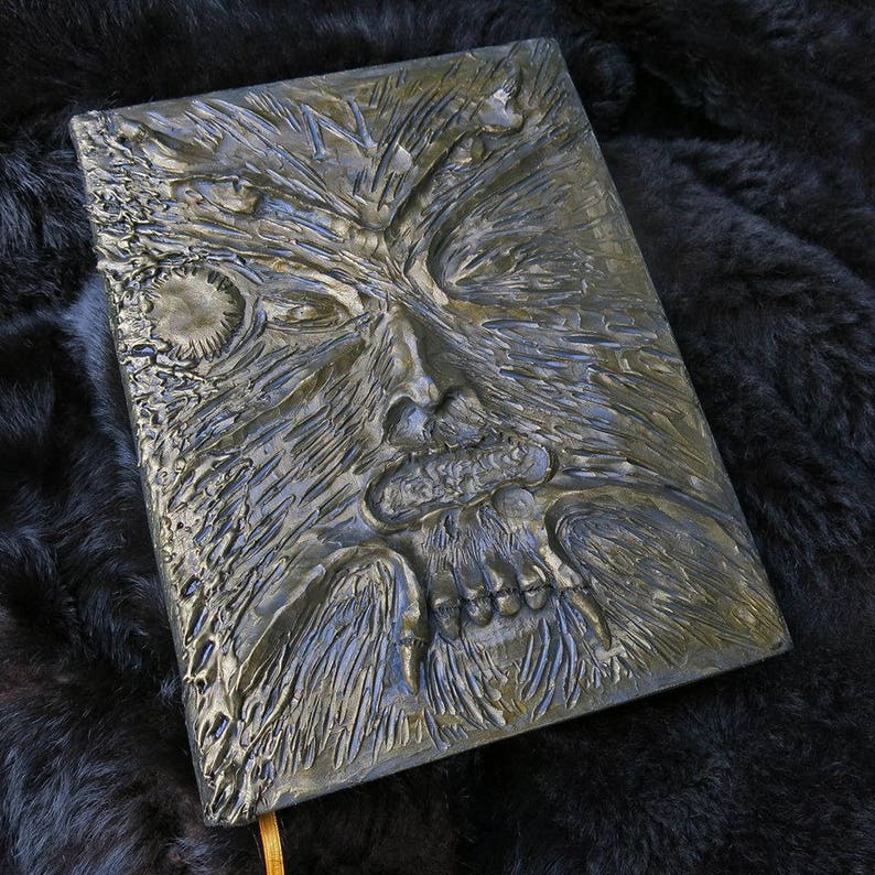 Puede incluir: Un diario de cuero gris oscuro con un dise&ntilde;o en relieve detallado de una cara grotesca con barba y dientes. La cara parece estar hecha de madera.