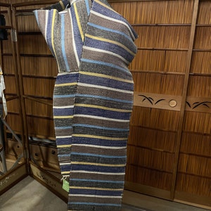May include: A striped scarf in shades of brown, blue, yellow, and gray is draped over a mannequin. The scarf has a textured appearance with horizontal stripes. The background includes a wooden screen and a concrete floor.