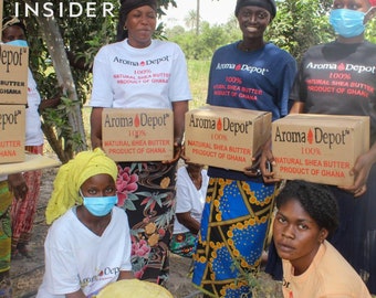 May include: A group of people holding boxes of 100% natural shea butter from Ghana, labeled "Aroma Depot". The boxes are brown with white text and a red logo. The people are smiling and holding the boxes in front of them. There are two large, round, brown objects in the foreground.