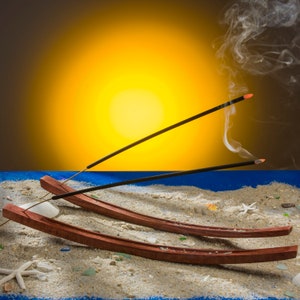 May include: Two curved, dark brown wooden incense holders resting on a sandy surface. Each holder contains a lit black incense stick with a glowing orange tip, emitting thin wisps of white smoke that curl upward against a bright yellow background.