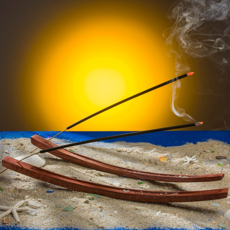 May include: Two curved, dark brown wooden incense holders resting on a sandy surface. Each holder contains a lit black incense stick with a glowing orange tip, emitting thin wisps of white smoke that curl upward against a bright yellow background.