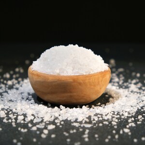 May include: A wooden bowl filled with white sea salt, with more salt scattered around the bowl on a black surface.