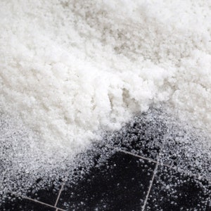 May include: A close-up of white salt crystals scattered on a black tiled surface. The salt is piled high and creates a textured, grainy effect.