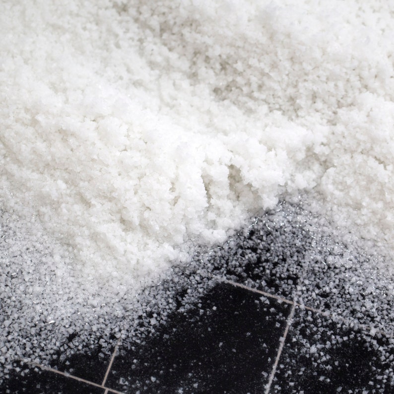 May include: A close-up of white salt crystals scattered on a black tiled surface. The salt is piled high and creates a textured, grainy effect.