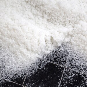 May include: A close-up of white salt crystals scattered on a black tiled surface. The salt is piled high and appears to be coarse.