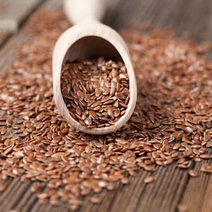 May include: A wooden scoop filled with brown flax seeds, spilling onto a wooden surface.