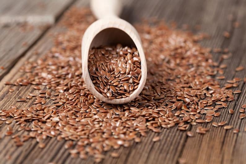 May include: A wooden scoop filled with brown flax seeds, spilling onto a wooden surface.