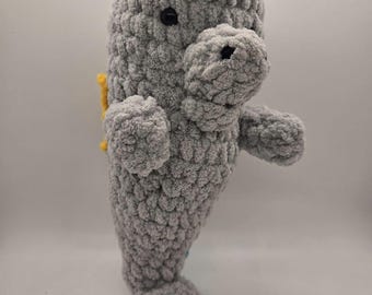 May include: A handmade grey crochet manatee plush toy with a pink flower on its head and yellow accents. The plush has small black eyes and a textured, soft appearance. The manatee is standing upright, showcasing its flippers and tail.