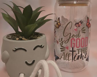 May include: A clear glass jar with a wooden lid and a straw. The jar features a design of butterflies and the text "God is good all the time". The jar is next to a grey ceramic planter with a smiley face and two white rope legs.