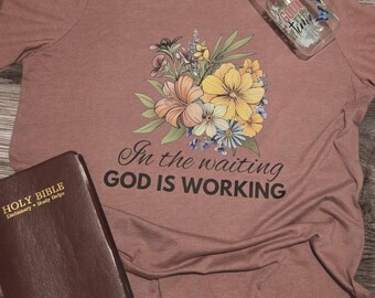 May include: A mauve t-shirt with a floral design and the text "In the waiting GOD IS WORKING". A brown leather-bound Holy Bible is in the bottom left corner of the image.