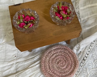May include: Two small, clear, scalloped-edge glass bowls filled with miniature pink ribbon roses and green ribbon leaves, resting on a rectangular wooden stool next to a round, dusty pink, hand-knitted wool coaster with a spiral texture.