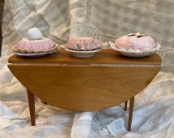May include: Three miniature desserts on small white plates resting on a wooden drop-leaf table. The left dessert is pink with a white dollop, the centre is a textured pink mould, and the right is a pink frosted treat with a small flower detail.