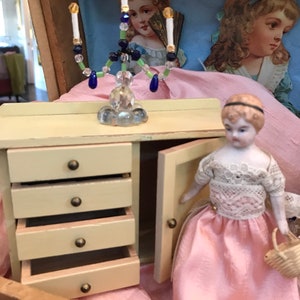 May include: A miniature cream-colored wooden chest of drawers with four drawers and a side cabinet door, a beaded candelabra with blue and green accents, and a small doll wearing a white lace top and a pink skirt holding a woven basket.