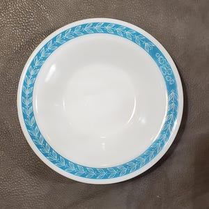 May include: White ceramic bowl with a turquoise leaf and floral pattern around the rim. The bowl has a simple, clean design, with a wide, shallow shape. The turquoise border provides a pop of color against the white background.