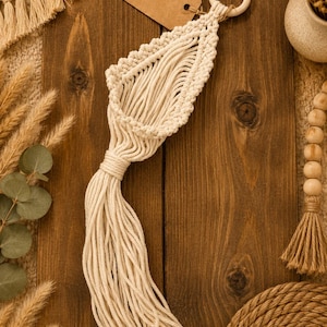May include: A cream-coloured macrame wall hanging featuring a leaf-shaped woven pocket, a wooden ring at the top, and a long fringe at the bottom. A brown rectangular tag attached to the ring displays the text THE WANDERING THREAD.