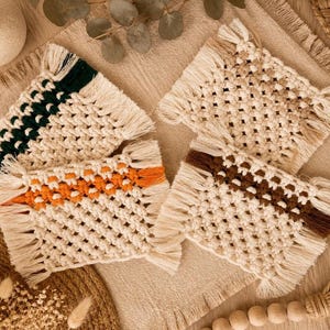 May include: Four rectangular macrame coasters made of off-white cotton cord. Each coaster features a central horizontal band of coloured knots in dark green, bright orange, and dark brown, with one coaster remaining entirely off-white.