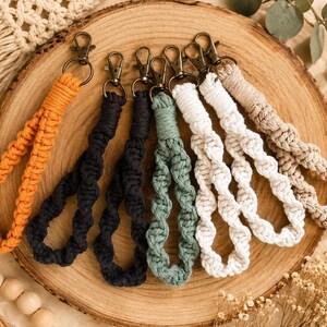 May include: A collection of seven macrame wristlet keychains featuring knotted cord in orange, black, dark gray, sage green, white, and beige. Each keychain is finished with a bronze-toned metal lobster clasp attached to the looped cord.