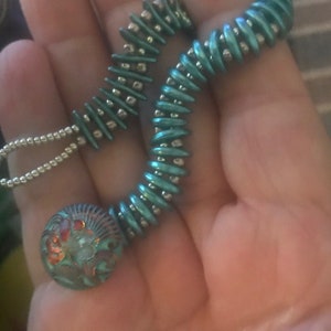 May include: A teal and silver beaded necklace with a round, multi-colored glass pendant. The pendant has a floral design with red, orange, and green accents.