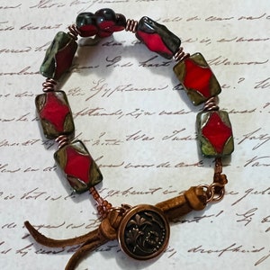 May include: A red and green glass bead bracelet with a copper clasp and a leather tassel. The bracelet has a floral design on the clasp.