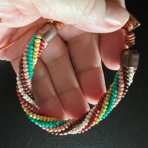 May include: A multi-colored beaded bracelet with brown leather ties and copper accents. The bracelet is made of small, round beads in shades of pink, white, brown, yellow, and turquoise.