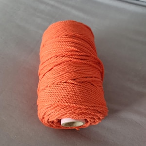 May include: A spool of bright orange macrame cord. The cord is tightly wound around a cardboard tube. The colour is a vibrant, solid orange, and the texture of the cord is clearly visible.
