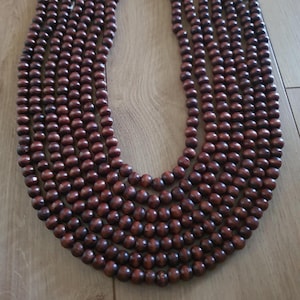 May include: A multi-strand necklace composed of dark brown wooden beads. The beads are round and evenly sized, creating a textured, layered effect. The necklace is displayed on a light brown wooden surface.