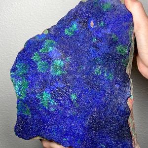 Pode incluir: Um grande espécime de azurite, de forma irregular. A pedra apresenta um tom azul profundo com manchas de verde vibrante. As bordas são marrons avermelhadas. O mineral é segurado pelas mãos de uma pessoa.
