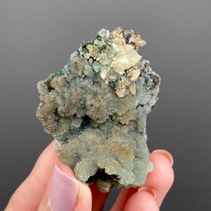 May include: A close-up of a mineral specimen featuring a cluster of pale green, crystalline formations. The specimen has a rough, textured surface with patches of light brown and beige crystals. The background is a solid gray.