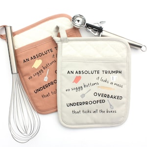 May include: Two square quilted oven mitts in dusty rose and cream colors, featuring printed text including "AN ABSOLUTE TRIUMPH" and "no soggy bottoms," accompanied by a stainless steel wire whisk and a metal ice cream scoop for baking.