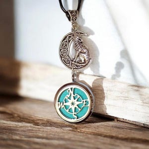 May include: Silver pendant necklace with a howling wolf on a crescent moon. Beneath, a round pendant with a teal background and a compass rose design. The necklace is suspended from a black cord, set against a wooden surface.