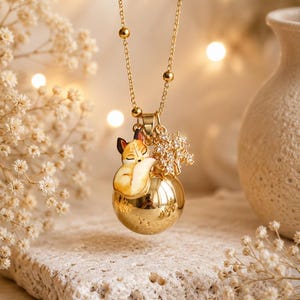 Puede incluir: Un collar de tono dorado con una cadena delicada de pequeñas cuentas, un colgante esférico pulido, un dije de zorro esmaltado en naranja y blanco, y un pequeño dije en forma de árbol incrustado con cristales transparentes brillantes.