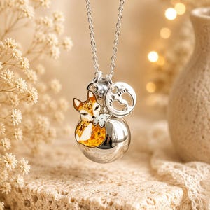 Op de afbeelding: Een zilverkleurige ketting met een grote, gladde, bolvormige hanger. Aan het oogje hangen twee bedels: een kleurrijke emaille vos met oranje, witte en zwarte details, en een kleine ronde bedel met de afdrukken van babyvoetjes.