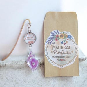 Peut inclure: Un marque-page en métal doré avec une breloque ronde portant l'inscription Merci Maitresse et un pendentif en verre violet en forme de coquillage, accompagné d'une enveloppe en papier kraft avec un autocollant Maitresse Parfaite Merci pour cette année.