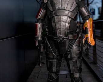 Könnte beinhalten: Eine Person in einem grauen und schwarzen futuristischen Anzug mit einem leuchtend orangefarbenen Arm-Anbauteil. Der Anzug trägt die Buchstaben "N7" auf der Brust.