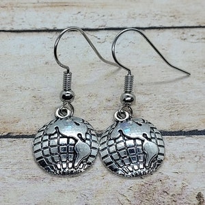 May include: A pair of silver-colored dangle earrings. Each earring features a globe charm with a detailed map design. The earrings have a hook closure and are displayed against a light wood grain background.