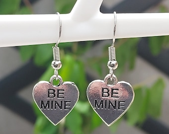 May include: Silver-toned heart-shaped earrings. Each earring features the words "BE MINE" in a bold, black font. The earrings hang from a silver hook and are displayed against a blurred green background.