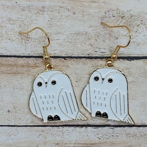 May include: A pair of gold-tone metal earrings featuring white owl pendants with black eyes and feet. The owls have gold outlines and are hanging from gold-colored ear wires. The earrings are a whimsical accessory.