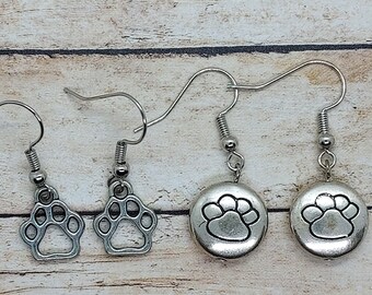 May include: Four pairs of silver-toned earrings featuring paw print designs. Two pairs have open paw prints, and two pairs have paw prints on round discs. The earrings hang from silver-colored fish hook ear wires.