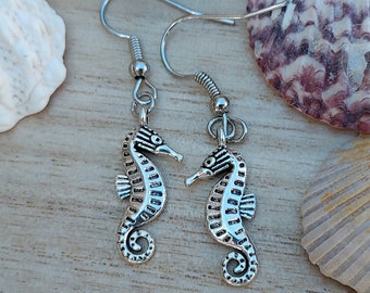 May include: A pair of silver-colored seahorse earrings. Each earring features a detailed seahorse charm suspended from a silver-tone ear wire. The seahorses have textured bodies and curled tails, evoking a nautical theme.